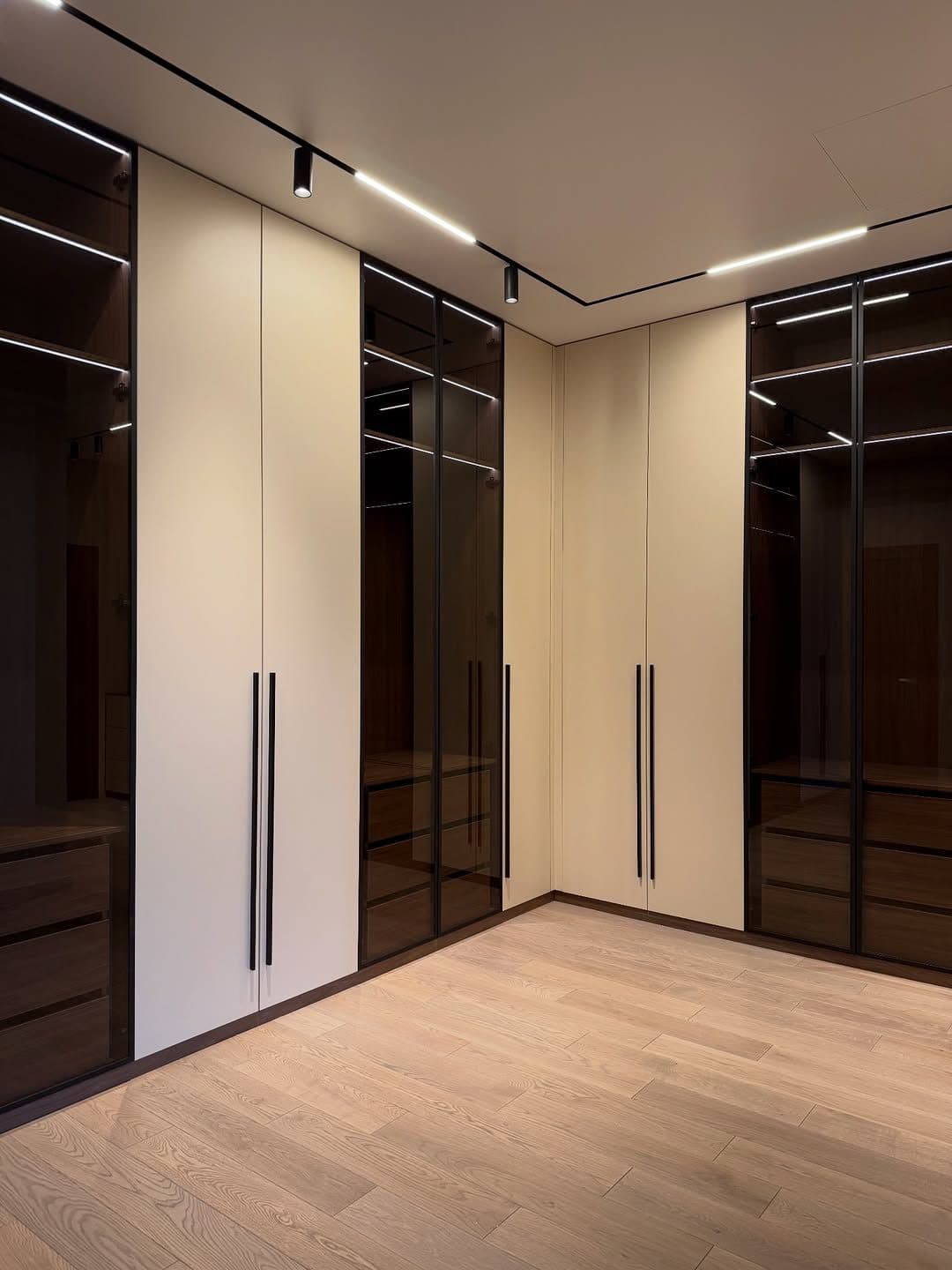 Walk-in dressing room with smoked-glass wardrobe doors, cream panels and track lighting by Royalwoods Interiors, Nairobi