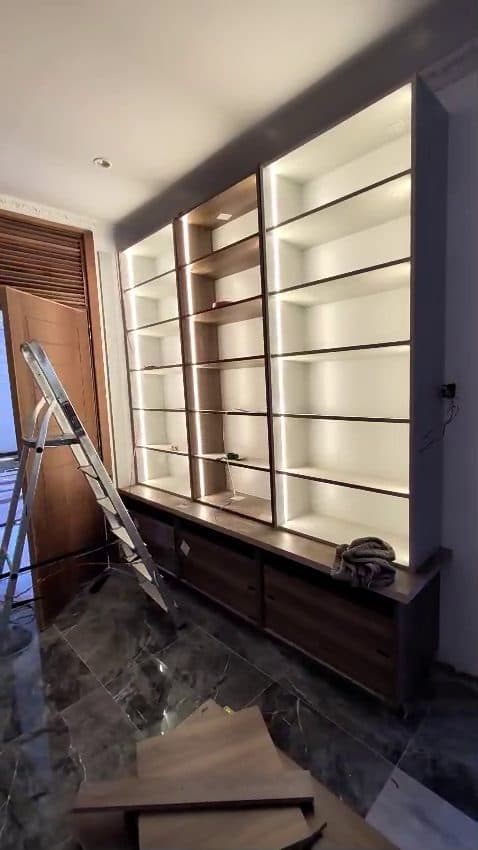 A full-height display wall with an LED channel in every shelf, set against dark timber and marble. — Royalwoods Interiors, Nairobi